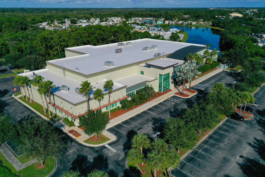 Aerial view of the LactaLogics processing facility in Port St. Lucie, Florida, a large pale building with a flat roof, surrounded by palm trees, a parking lot, and a lake in the distance.