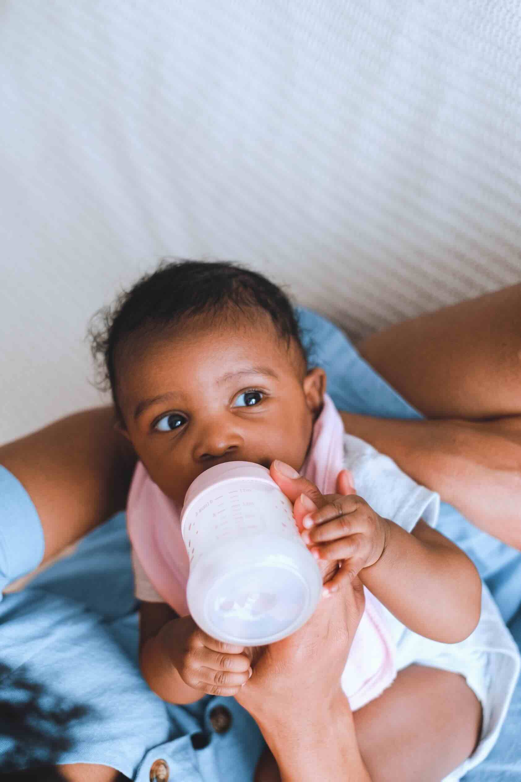 A baby with dark curly hair looking up at the camera while drinking from a bottle, held by an adult in a light blue top.