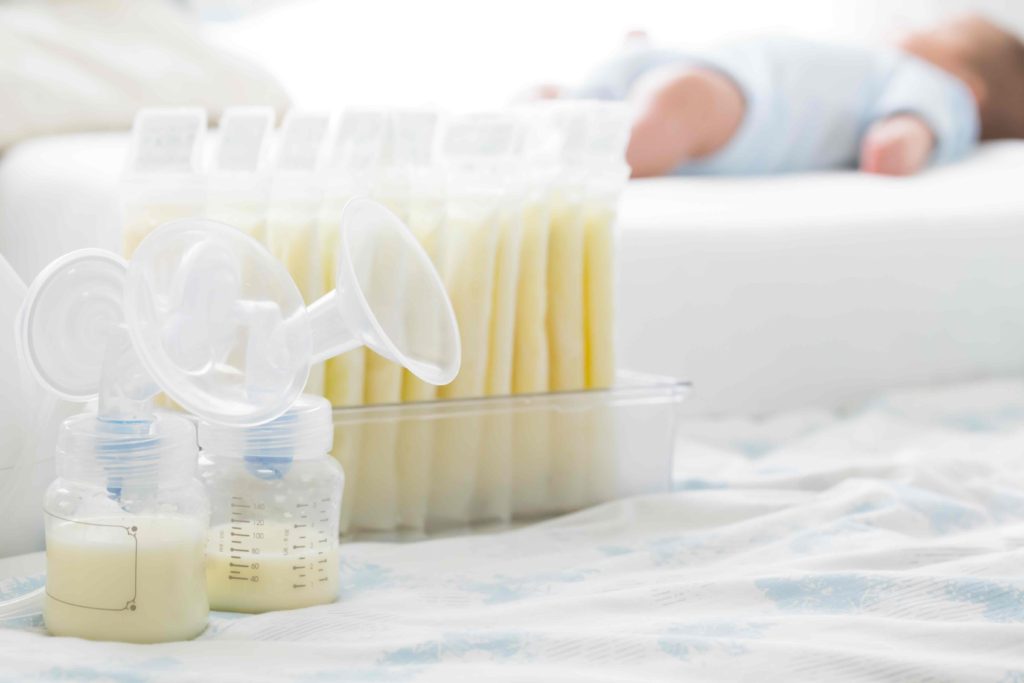 Rows of frozen breast milk storage bags standing upright in a clear tray, with a breast pump flange and a partly filled collection bottle in front, and a baby sleeping out of focus in the background.